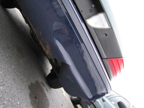 1991 Ford LTD Crown Victoria - Close-up of a blue car's rear quarter panel and taillight, revealing rust and damage. Another vehicle is partially visible.