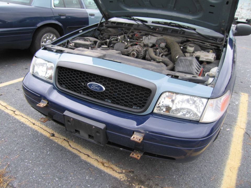 Front of a blue Ford Crown Victoria with hood open, showing engine. Another blue car is parked behind it
