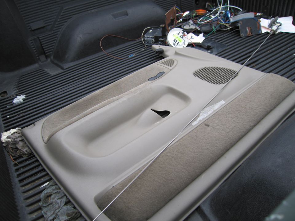 Car door panel with speaker grille and armrest, lying in a truck bed with wiring and parts in the background