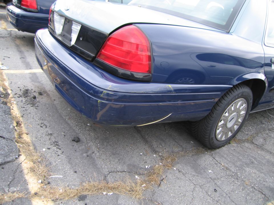 Blue sedan's rear bumper and taillight, showing scuffs and scratches. Another blue sedan is partially visible behind it