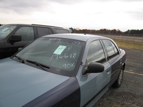 1991 Ford LTD Crown Victoria - A two-tone Ford Crown Victoria has handwritten notes on its windshield, parked next to a dark SUV under an overcast sky.