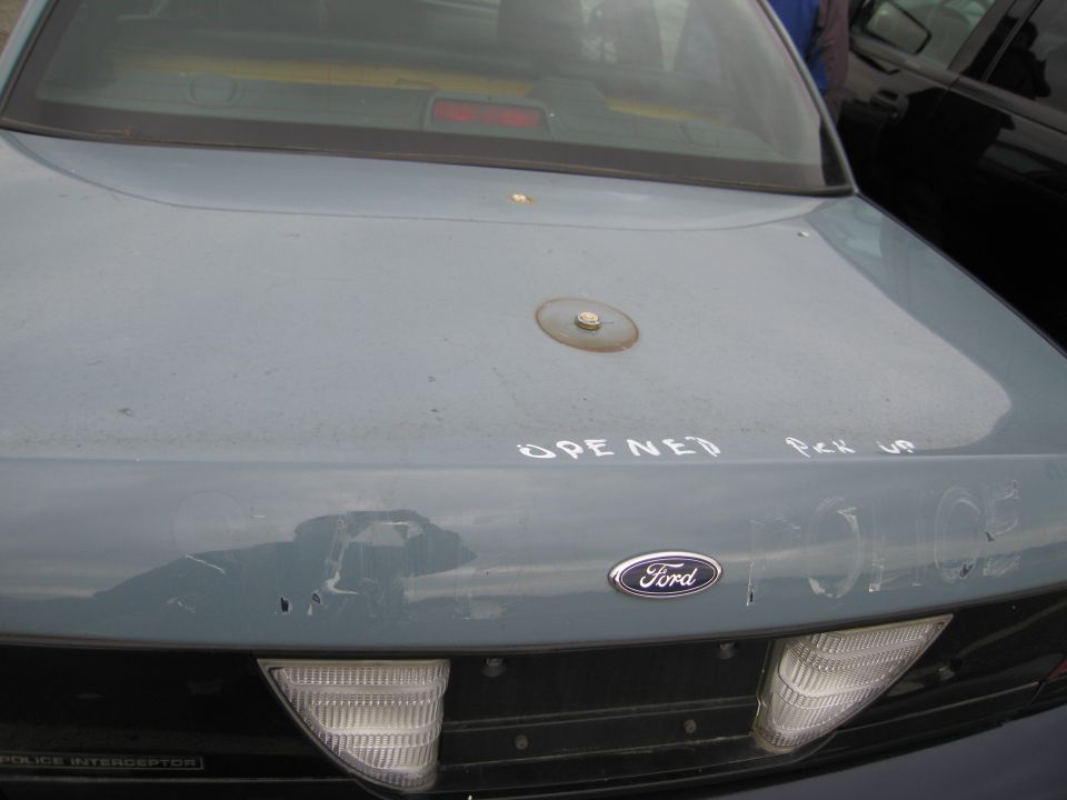 Rear of a light blue Ford sedan with "OPENED Pick UP" and "POLICE" written on the trunk