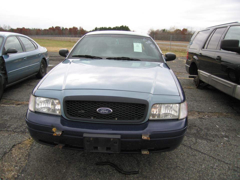 Front view of a light blue and dark blue Ford Crown Victoria. Another car is visible to the left, and a black SUV is to the right
