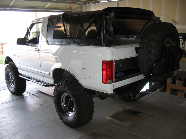 White lifted Ford Bronco with black soft top and large off-road tires, rear 3/4 view in a garage