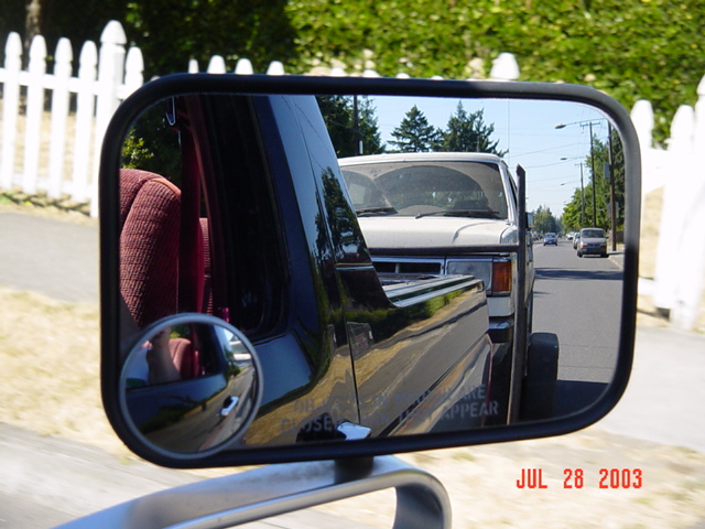 Side mirror reflection showing white truck being towed behind black vehicle, red interior, dated 2003