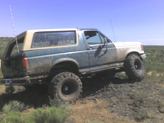 Mud-splattered Ford Bronco, two-tone green and white, lifted with off-road tires, viewed from the side on uneven terrain