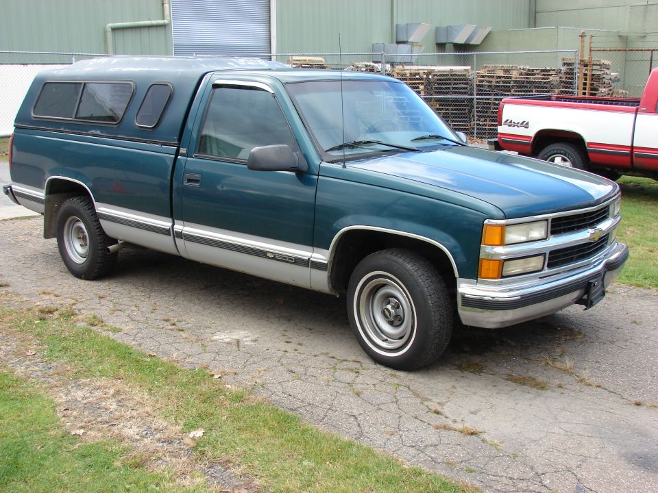Green pickup truck with a silver stripe and a camper shell parked on cracked asphalt. Another red and white truck is visible in the background