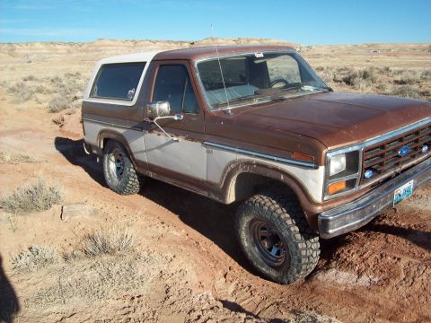 1985 Ford Bronco - This two-tone Ford Bronco is shown off-roading on a dusty trail, its tires gripping the uneven terrain.