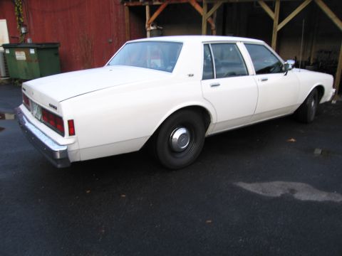 1991 Ford LTD Crown Victoria - A white sedan is parked outside a building with a red wall and wooden beams overhead.