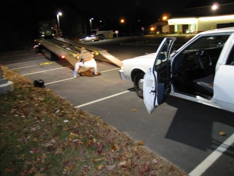 1991 Ford LTD Crown Victoria - A white sedan is being loaded onto a tow truck at night, with its driver's door ajar.