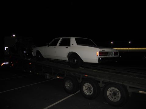1991 Ford LTD Crown Victoria - A white sedan is transported on a car carrier at night, showing its rear profile. The vehicle's styling suggests it is from the 1980s.