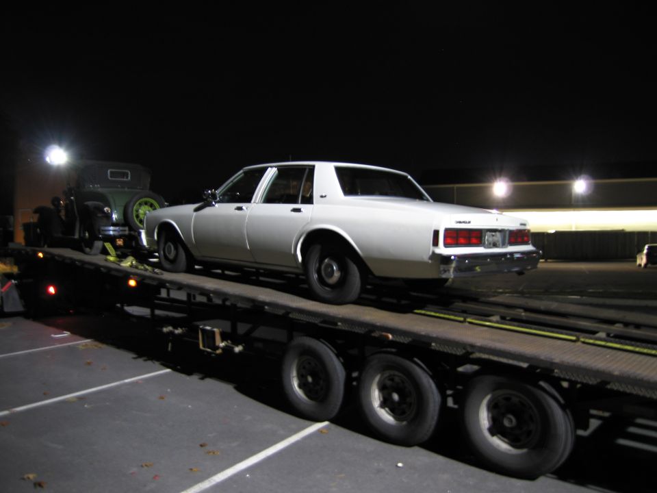 White sedan on a car carrier trailer at night, with a green vintage car also on the trailer