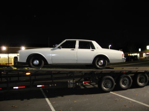 1991 Ford LTD Crown Victoria - A white sedan is transported on a trailer at night, with lights from a nearby building in the background.