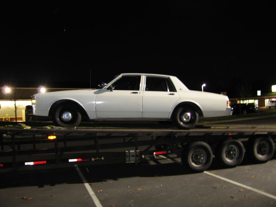 White sedan on a trailer at night. The car has a boxy shape and steel wheels