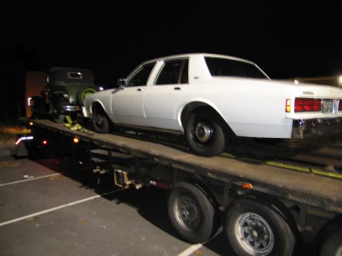 1991 Ford LTD Crown Victoria - A white sedan is transported on a car hauler trailer at night, with another vehicle visible behind it.