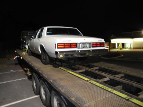 1991 Ford LTD Crown Victoria - A white sedan is being transported on a flatbed trailer at night, with a building visible in the background.