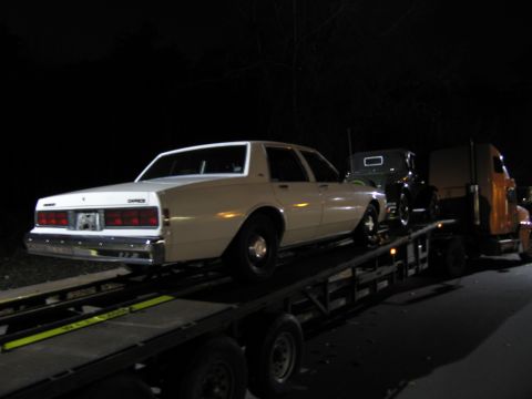 1991 Ford LTD Crown Victoria - A white sedan is loaded onto a car carrier, with another vehicle visible behind it.