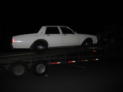1991 Ford LTD Crown Victoria - A white sedan is loaded onto a car hauler at night, with another vehicle partially visible behind it.