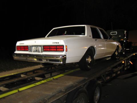 1991 Ford LTD Crown Victoria - A white sedan is loaded onto a car carrier, with another vehicle partially visible behind it in the darkness.