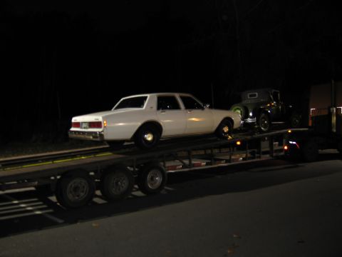 1991 Ford LTD Crown Victoria - Two cars are transported on a trailer at night, one a white sedan and the other a vintage model.