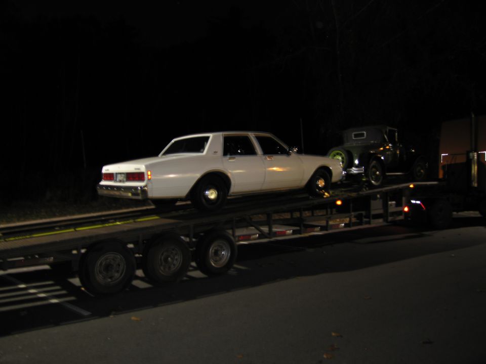 White sedan on a trailer at night, side view. A dark vintage car is behind it