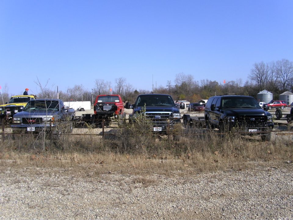 Four trucks and SUVs parked in a lot, front view. Black Ford F-250, black Ford Excursion, red truck chassis, and a blue GMC pickup