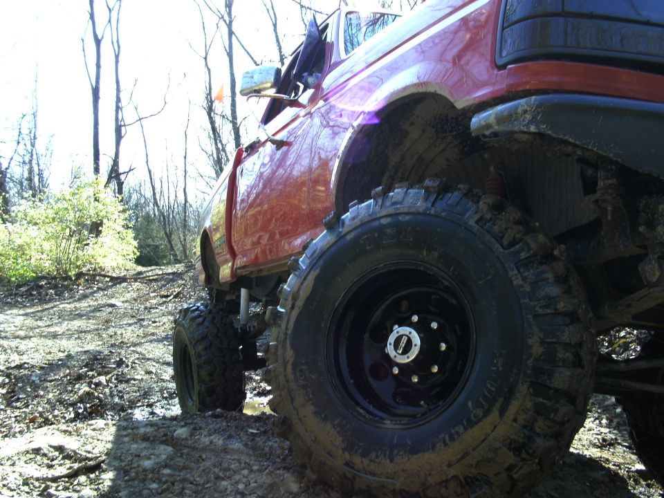 Red pickup truck with large off-road tires on a muddy, wooded trail. Front fender and door visible