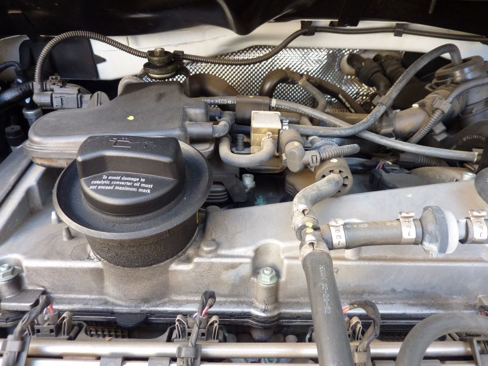 Engine bay close-up of aluminum valve cover, oil filler cap with warning text, and hoses