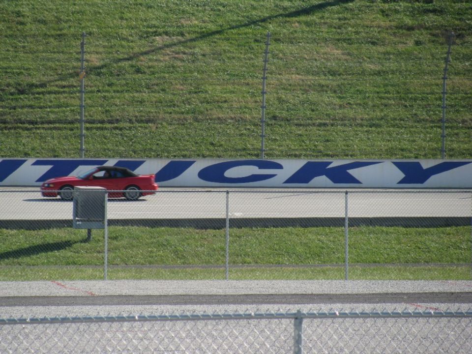 Red coupe with black top on racetrack, with 'Kentucky' signage