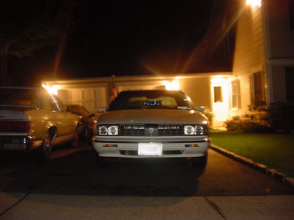 Nighttime front view of white Oldsmobile sedan in a driveway, headlights on, house lit behind