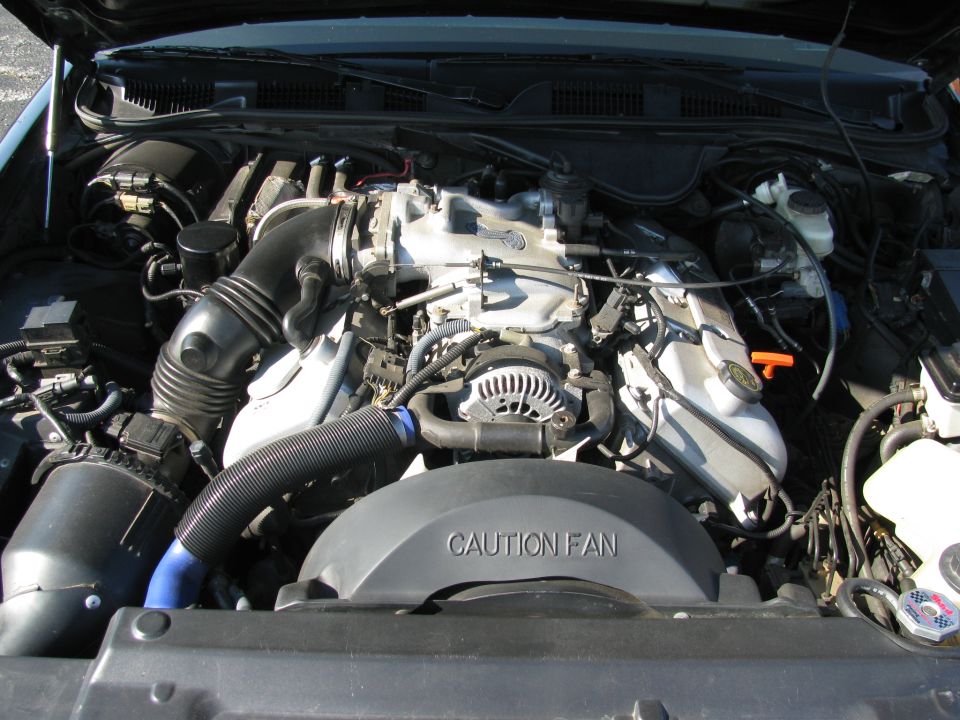 Engine bay with V8 engine components, air intake, alternator, and fan shroud labeled 'CAUTION FAN'