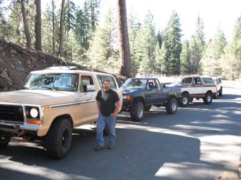 1991 Ford Bronco - A line of off-road vehicles, including a tan 1978 Ford Bronco, parked on a paved road in a wooded area.
