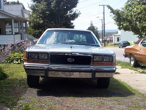 1991 Ford LTD Crown Victoria - Head-on front view of a light silver Ford Crown Victoria sedan parked on a gravel driveway beside a white house, with hood ornament and For…