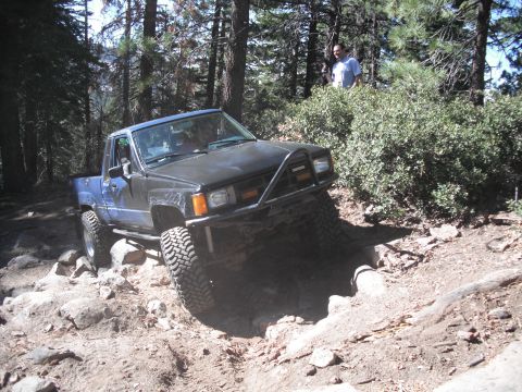 1991 Ford Bronco - A Toyota pickup truck with a custom bumper navigates a challenging rocky terrain on a wooded trail.