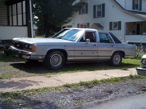 1991 Ford LTD Crown Victoria - A silver 1980s-90s Ford full-size sedan parked at an angle on a concrete driveway, shown from the front-left 3/4 view with a white two-stor…
