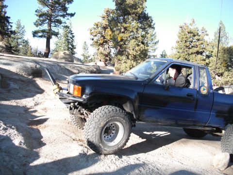 1991 Ford Bronco - Pickup truck navigating a rocky, off-road trail with trees and boulders in the background.