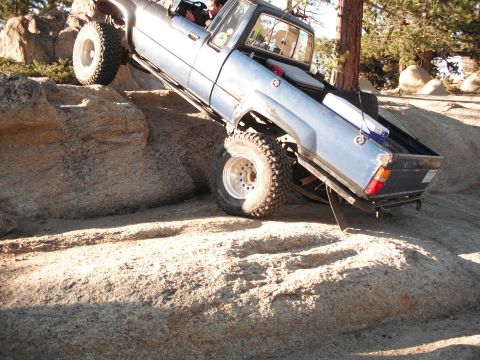1991 Ford Bronco - A blue pickup truck navigates a rocky off-road trail, with its tires angled on the uneven terrain.