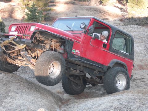 1991 Ford Bronco - A red Jeep Wrangler navigates a rocky trail, with one wheel lifted off the ground.