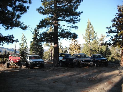1991 Ford Bronco - Multiple off-road vehicles are parked on a dirt lot under trees with mountains visible in the distance.