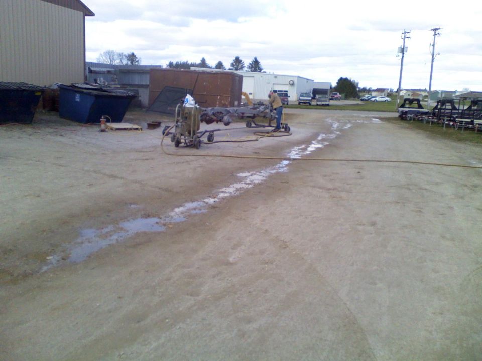 Vehicle frame on a stand with a sandblaster hose and operator nearby