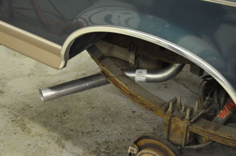Close-up of a vehicle's rear axle, leaf springs, and exhaust pipe. A white sticker with text is visible on the exhaust pipe
