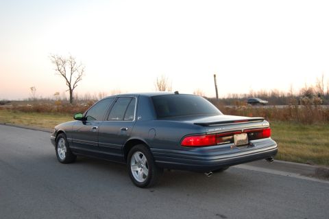 1985 Mercury Grand Marquis - A blue sedan is parked on the side of a road with a tree in the background.