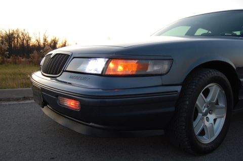 1985 Mercury Grand Marquis - The front end of a Mercury Grand Marquis is shown with the headlights and grille visible against a sunset sky.