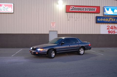 1985 Mercury Grand Marquis - A dark blue sedan, possibly a 1998 model, is parked in a lot outside a building with various tire brand signs.