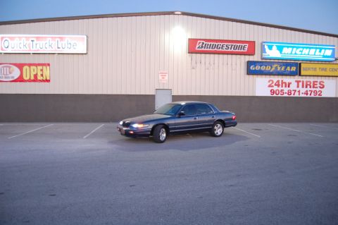 1985 Mercury Grand Marquis - A dark blue sedan is parked in a lot outside a building with various tire company advertisements on the wall.