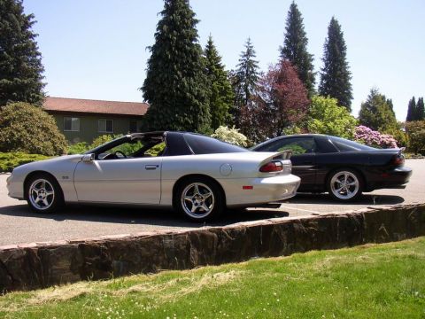 1988 Ford Bronco - Two Chevrolet Camaros, a silver convertible and a black coupe, are parked next to each other on a sunny day.