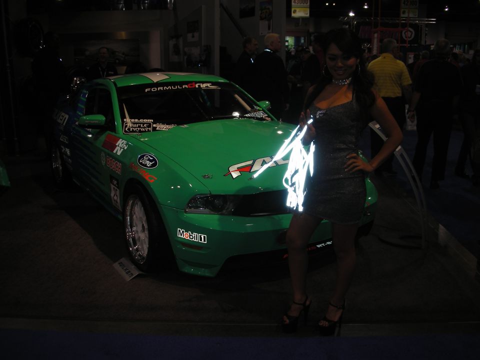 Green Ford Mustang race car with Formula Drift decals, parked indoors next to a woman