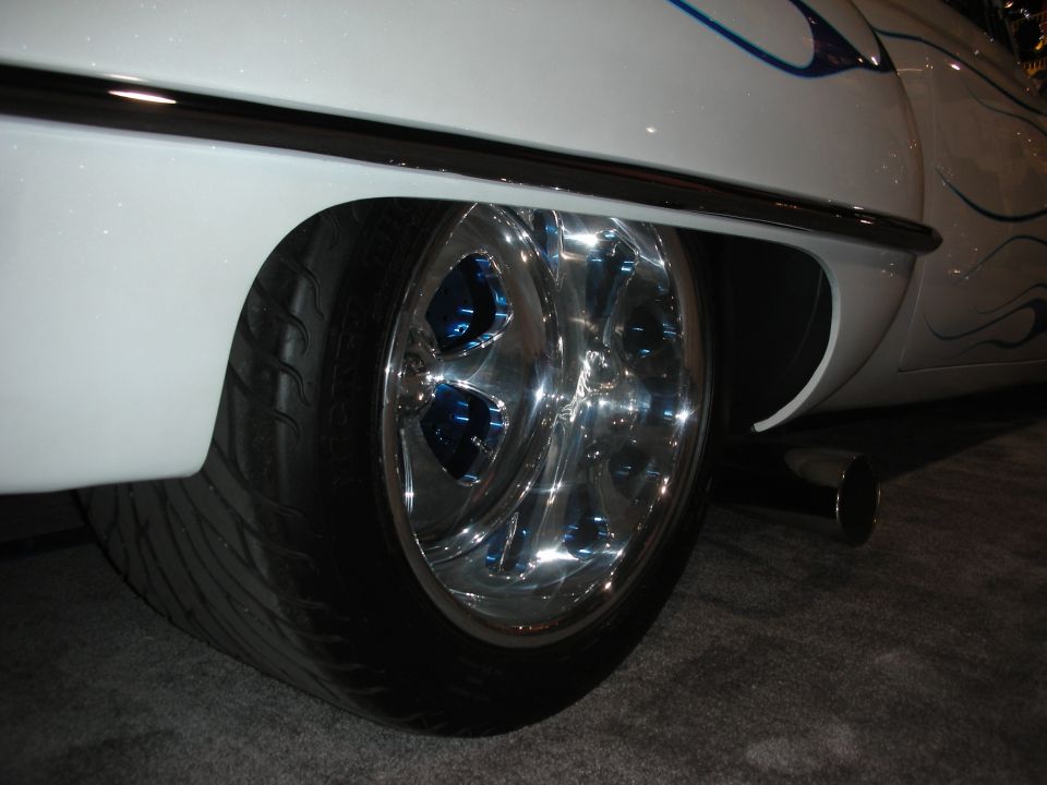 Close-up of a custom car's rear wheel with chrome rims and blue accents, white body with blue flame graphics, and a visible exhaust pipe