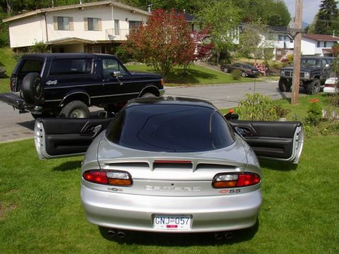 1988 Ford Bronco - Rear of a silver Chevrolet Camaro SS with both doors open parked on grass, a black lifted Ford Bronco behind it, plate GNJ 057.