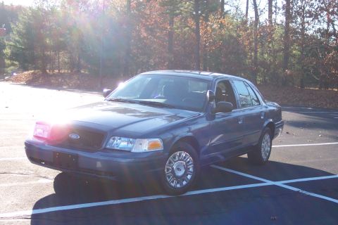 2001 Ford Crown Victoria - A dark blue 2001 Ford Crown Victoria is parked in a lot, with trees and autumn foliage in the background.